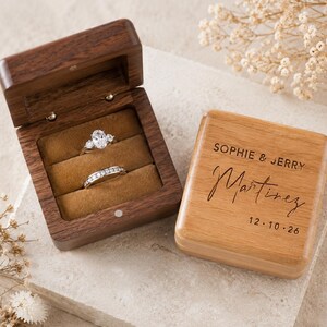 May include: A dark wood ring box with a hinged lid, containing two silver-toned rings with clear stones. Beside it, a light wood lid is engraved with the text SOPHIE & JERRY Martinez 12.10.26. The interior of the box is lined with brown velvet.