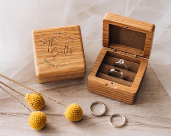 May include: A square wooden ring box with a hinged lid engraved with the text James & Katie, The Smiths, September 17, 2020. The open box features a brown velvet interior holding two silver rings, with two additional silver rings placed nearby.