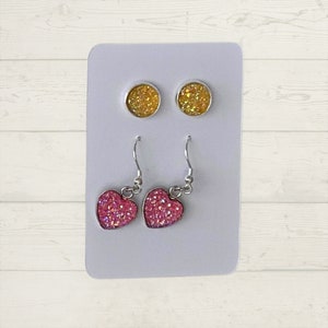 May include: A set of earrings featuring two pairs of stud earrings and two pairs of dangle earrings. The stud earrings are round and have a gold glitter finish. The dangle earrings are heart-shaped and have a pink glitter finish.