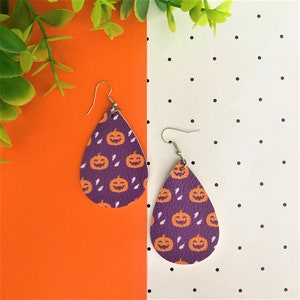May include: A pair of purple teardrop-shaped earrings with a pumpkin pattern. The earrings are made of leather and have silver hooks.