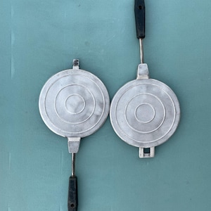 May include: Two circular silver-coloured metal waffle irons featuring concentric ring patterns on their surfaces. Each iron is equipped with a long metal rod and a black handle. The irons are positioned side-by-side, showcasing their flat, textured cooking plates and sturdy construction for heat-based food preparation.
