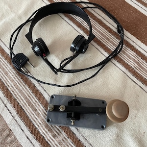 May include: A black wired headset with a two-prong plug and a telegraph key featuring a rectangular gray base, a black metal lever, and a round tan knob. The telegraph key has visible metal screws and a mechanical design for manual signal transmission.