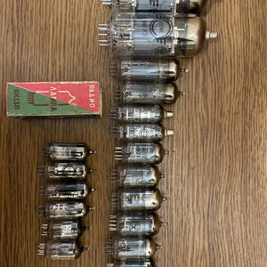 May include: A selection of vintage vacuum tubes, featuring glass casings and metal pins, displayed on a wooden surface. A small box with Cyrillic text and a green and red design is also visible. The tubes are of various sizes and shapes, with internal components.