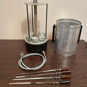 Op de afbeelding: Een elektrische rotisserie met een zwarte basis, zilveren accenten en een handvat. Inclusief een metalen kolen schoorsteen en verschillende metalen spiesen met houten en zwarte handvatten. De items staan op een donkerbruin houten oppervlak.