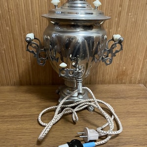 Può includere: Un samovar argentato lucido con dettagli ornati, manici bianchi e un rubinetto. Il cavo elettrico ha un motivo bianco e nero. Il samovar è su una superficie di legno.