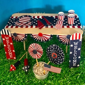 May include: A miniature patriotic table setting with red, white, and blue decorations. The table is decorated with a red, white, and blue tablecloth, paper plates, and cups. There are also two welcome signs with stars and stripes. The table is set on a green grass surface.