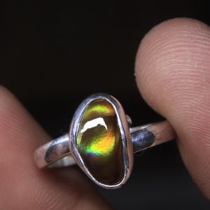 May include: A silver ring featuring a polished, oval-shaped gemstone with iridescent colors of yellow, green, and brown. The gemstone is set in a bezel setting, and the ring band is a simple, polished silver.
