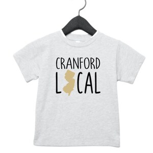 May include: A light gray t-shirt with black text that reads "Cranford Local" and a gold outline of the state of New Jersey.