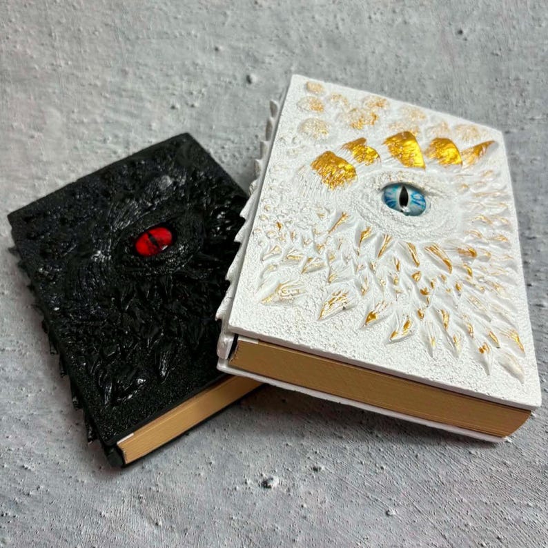 May include: Two rectangular journals with textured covers. The black journal features a single red eye, while the white journal has a blue eye surrounded by gold-accented scales. Both books have gold-colored page edges and a thick, rigid construction.
