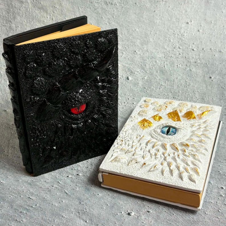 May include: Two hardcover books with textured, sculpted covers. The larger book is black with a prominent red eye at the center. The smaller book is white with gold accents and a blue eye at the center. Both books have tan pages visible at the edges.