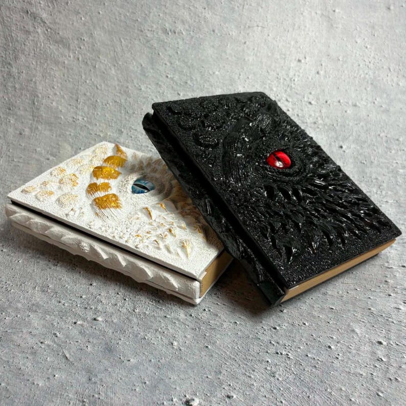 May include: Two rectangular journals with textured covers. One journal is black with a prominent red eye in the center. The other journal is white with gold-toned accents and a blue eye in the center. Both covers feature raised, scale-like details.