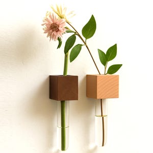 Könnte beinhalten: Zwei Wandvasen aus Holz, eine dunkelbraune und eine helle, jede mit einem durchsichtigen Glasröhrchen. Die linke Vase enthält eine rosa Blume, die rechte grüne Blätter. Minimalistische Blumendekoration.