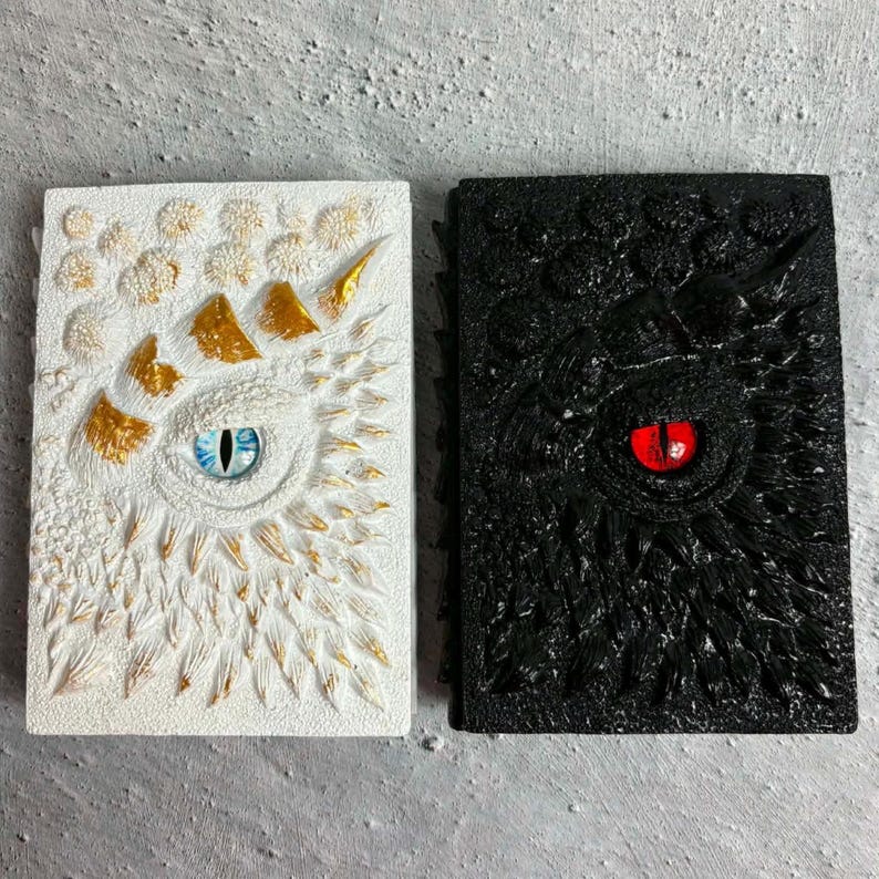 May include: Two rectangular journals with textured covers. The left journal is white with gold accents and a central blue eye. The right journal is black with a central red eye. Both covers feature raised, scale-like patterns and sculpted details.