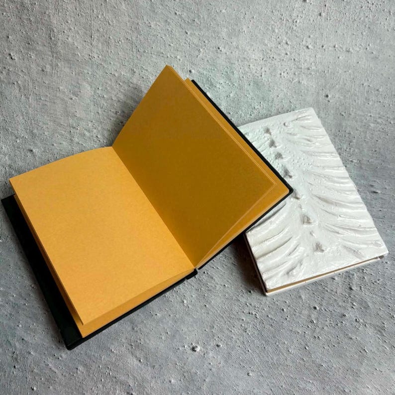 May include: An open notebook with blank, light brown pages and a black spine, positioned next to a closed notebook featuring a white, textured, raised relief cover with a repeating pattern of curved, organic lines across the surface.