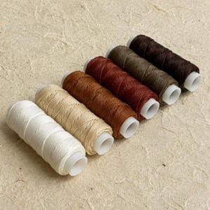 May include: Six spools of sewing thread arranged in a diagonal line, featuring a gradient of colours from white to beige, light brown, reddish-brown, olive green, and dark brown. Each spool has a white plastic core and tightly wound thread.