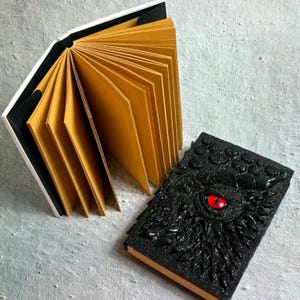 May include: A black journal with a textured, scaly cover featuring a prominent, singular red eye in the center. Beside it, an open book displays numerous thick, golden-yellow pages fanned out, revealing a black spine and white outer edges.