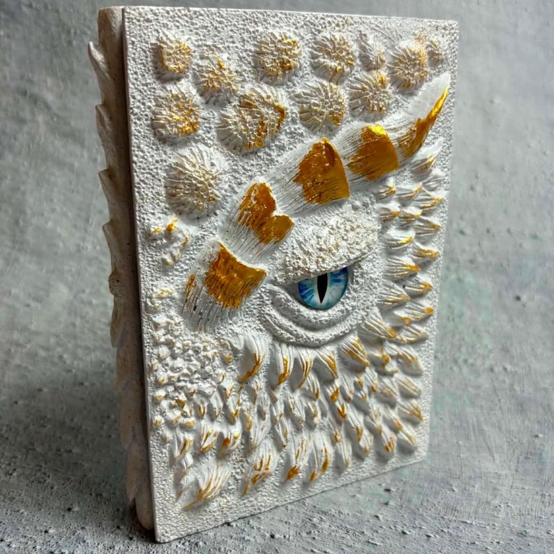May include: A rectangular journal with a textured white cover featuring a sculpted dragon eye with a blue iris and pupil. The surface is adorned with raised, scale-like patterns and circular shapes, accented with metallic gold paint highlights.