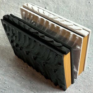 May include: Two rectangular objects with textured, raised surfaces resembling book covers. The front object is matte black with a thick, uneven texture, while the back object is white with a similar raised pattern. Both have visible tan page edges.