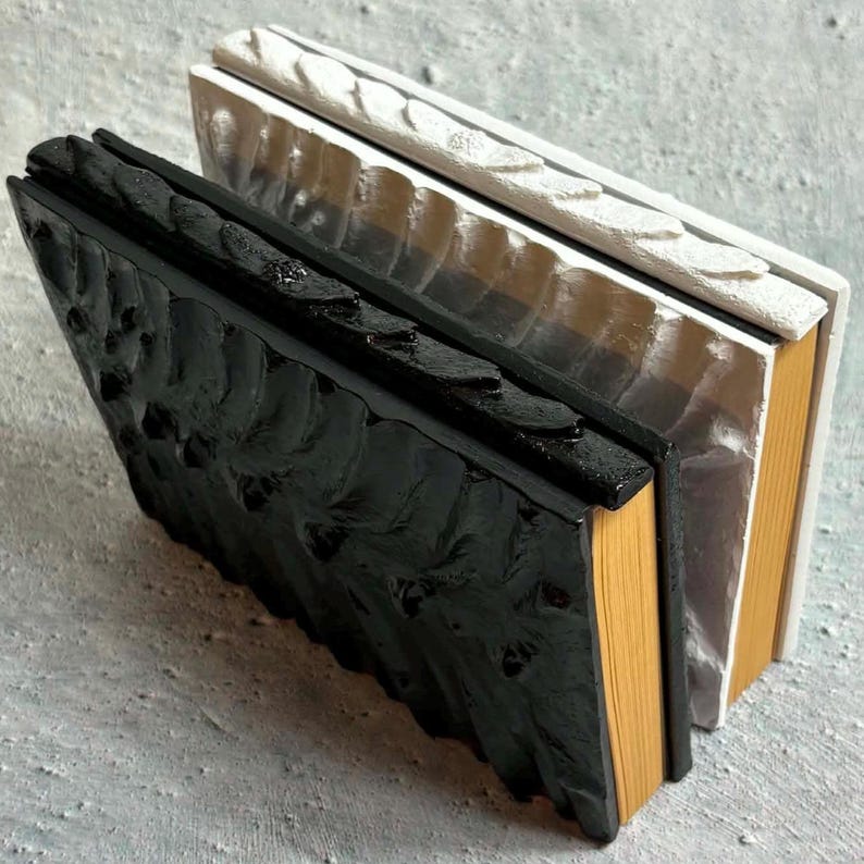 May include: Two rectangular objects with textured, raised surfaces resembling book covers. The front object is matte black with a thick, uneven texture, while the back object is white with a similar raised pattern. Both have visible tan page edges.