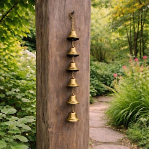 May include: A brass wind chime with six bells hanging from a chain. The bells are suspended from a wooden post in a garden setting. The background features lush greenery and a stone pathway.