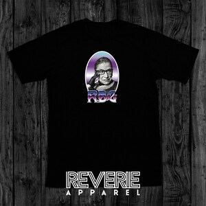 Op de afbeelding: Zwart T-shirt met een afbeelding van Ruth Bader Ginsburg in een paarse en blauwe ovaal met de tekst "RBG" in een zilveren lettertype. De tekst "REVERIE APPAREL" is in wit onderaan het shirt gedrukt.