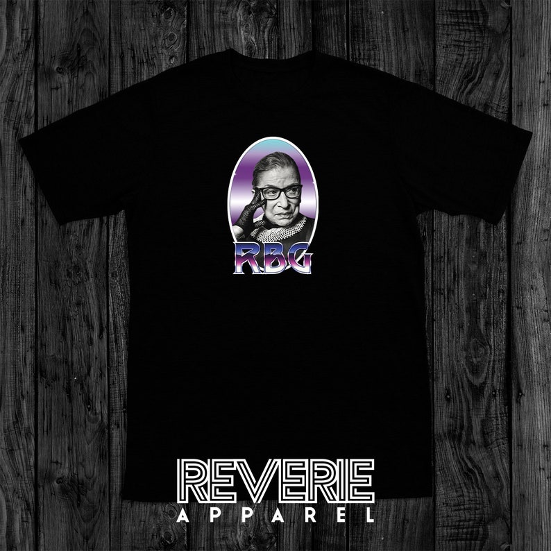 Op de afbeelding: Zwart T-shirt met een afbeelding van Ruth Bader Ginsburg in een paarse en blauwe ovaal met de tekst "RBG" in een zilveren lettertype. De tekst "REVERIE APPAREL" is in wit onderaan het shirt gedrukt.