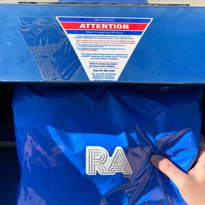 Op de afbeelding: Een blauwe USPS-brievenbus met een bord met de tekst "AANDACHT Postverzamelbeleid" en een witte sticker met de tekst "RA".
