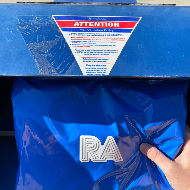 Op de afbeelding: Een blauwe USPS-brievenbus met een bord met de tekst "AANDACHT Postverzamelbeleid" en een witte sticker met de tekst "RA".