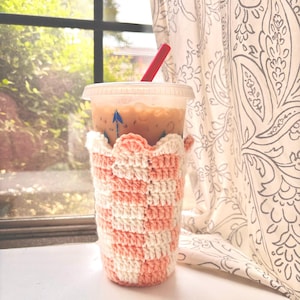 May include: A pink and white crocheted cup cozy with a scalloped edge. The cozy is on a tall iced coffee cup with a red straw.