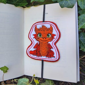 Könnte beinhalten: Handgefertigter Lesezeichen mit einem Cartoon-Drachendesign. Der orangefarbene Drache mit grünen Augen ist auf einem weißen Hintergrund mit rotem Rand aufgenäht. Das Lesezeichen befindet sich in einem offenen Buch.