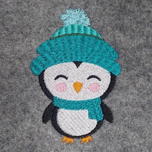 Op de afbeelding: Geborduurde pinguïn patch met een teal gebreide muts en sjaal. De pinguïn heeft een witte body, zwarte vleugels en een oranje snavel en poten. De achtergrond is grijs vilt.
