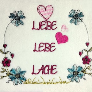 Könnte beinhalten: Gesticktes Design mit den deutschen Worten "Liebe, Lebe, Lache" (Liebe, Lebe, Lache) in Burgunderrot. Das Design umfasst blaue Blumen, rosa Herzen und einen grünen Grasrand.