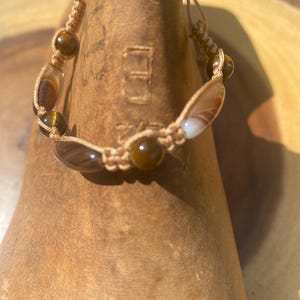 May include: A handmade bracelet featuring tiger's eye and agate stones, strung on a beige cord. The bracelet has a macrame design with alternating round and oval-shaped stones. The stones display shades of brown, tan, and cream.