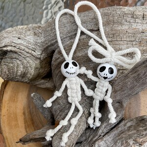 May include: Two handmade white macrame skeleton ornaments with black painted faces and attached loops for hanging. The skeletons are holding hands and are displayed on a piece of driftwood. These Halloween decorations are perfect for adding a spooky touch to any space.