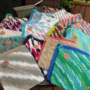 Può includere: Una collezione di colorati asciugamani di stoffa lavorati a maglia a mano. Gli asciugamani presentano vari motivi e colori, tra cui strisce e tinte unite. Ogni panno ha un anello per appenderlo e una piccola etichetta "Handmade with love."