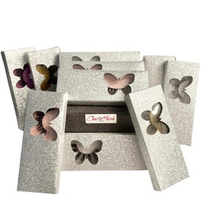 May include: A display of silver glitter eyelash boxes, each featuring a butterfly-shaped cutout. Some boxes are open, showcasing the eyelashes within. The label "Charis Made" is visible on one of the boxes.