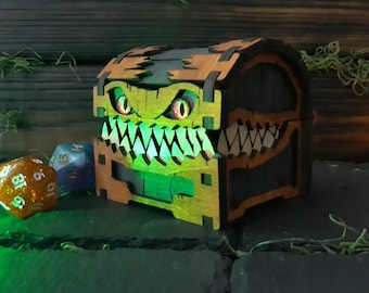 May include: A wooden treasure chest with a monster face design, featuring glowing green eyes and sharp teeth. The chest is brown and black, with a green interior. Dice are placed near the chest, and a small campfire is in the background.