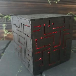 May include: A dark wooden cube with a circuit board design cut into its sides. The interior glows with a red light, creating a striking contrast. The cube sits on a dark, textured surface, suggesting a modern or tech-inspired theme.