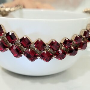 May include: A white teacup adorned with a band of square, ruby-red gemstones set in a gold-coloured metal. The gemstones are arranged in a repeating pattern around the cup's rim. The cup is set against a blurred background.