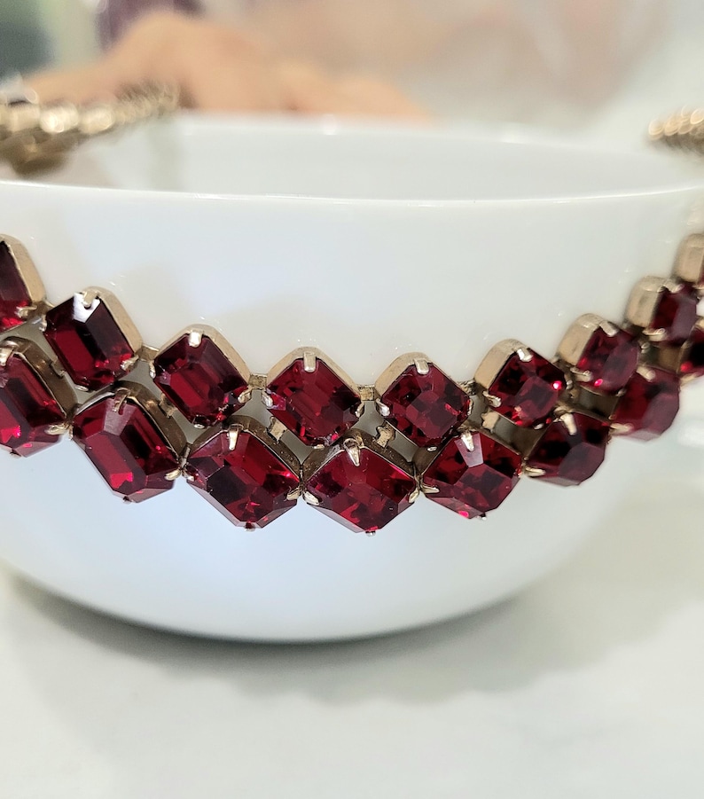 May include: A white teacup adorned with a band of square, ruby-red gemstones set in a gold-coloured metal. The gemstones are arranged in a repeating pattern around the cup's rim. The cup is set against a blurred background.