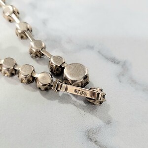 May include: Close-up of a silver necklace featuring round, faceted stones. The necklace has a delicate chain and a clasp with the brand name "WEISS" engraved. The stones are set in a prong setting.