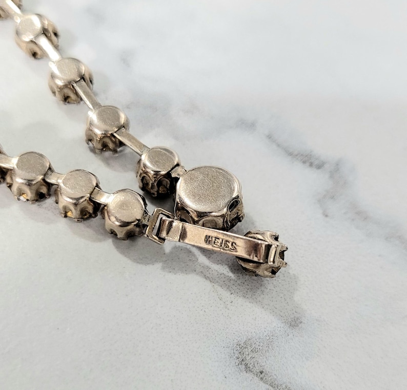 May include: Close-up of a silver necklace featuring round, faceted stones. The necklace has a delicate chain and a clasp with the brand name "WEISS" engraved. The stones are set in a prong setting.
