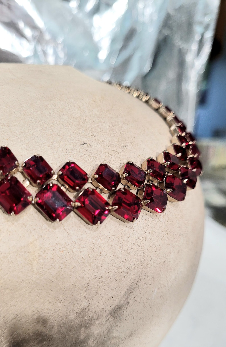 May include: A vintage necklace with two rows of rectangular, deep red gemstones set in gold-toned metal. The gemstones are closely set, creating a continuous band of colour. The necklace is displayed on a neutral-toned surface.