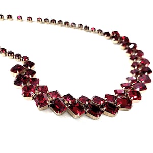May include: A vintage necklace with rectangular, ruby-red gemstones set in silver-toned metal. The necklace has a graduated design, with larger stones at the centre and smaller ones towards the clasp. The faceted stones catch the light, creating a sparkling effect.