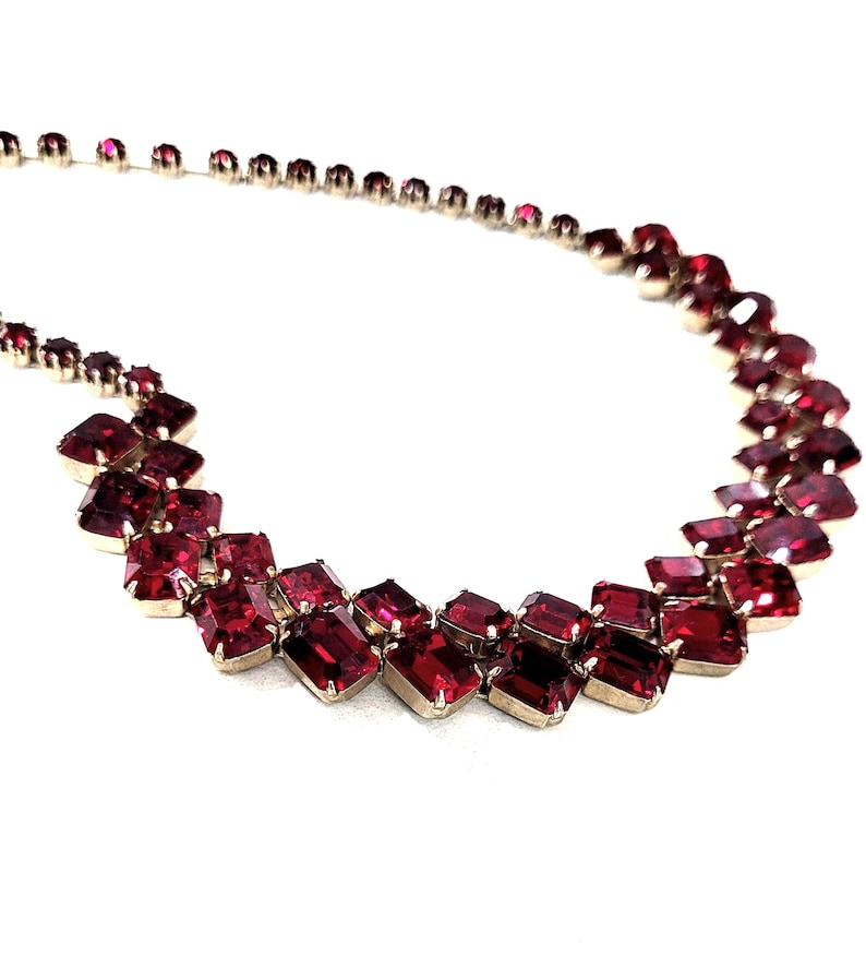May include: A vintage necklace with rectangular, ruby-red gemstones set in silver-toned metal. The necklace has a graduated design, with larger stones at the centre and smaller ones towards the clasp. The faceted stones catch the light, creating a sparkling effect.