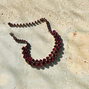 May include: A vintage necklace with rectangular, deep red gemstones set in gold-toned metal. The stones are arranged in a zig-zag pattern. The necklace is displayed on a neutral surface. The design is bold and eye-catching.