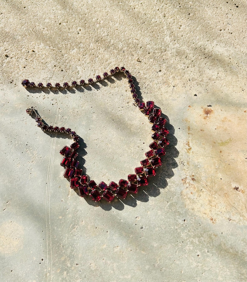 May include: A vintage necklace with rectangular, deep red gemstones set in gold-toned metal. The stones are arranged in a zig-zag pattern. The necklace is displayed on a neutral surface. The design is bold and eye-catching.