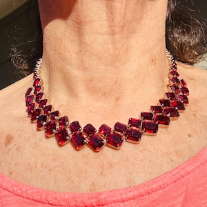May include: A gold-coloured necklace with a series of rectangular, deep red gemstones. The stones are set closely together, creating a bold design. The necklace is a statement piece, ideal for adding a touch of glamour to an outfit.
