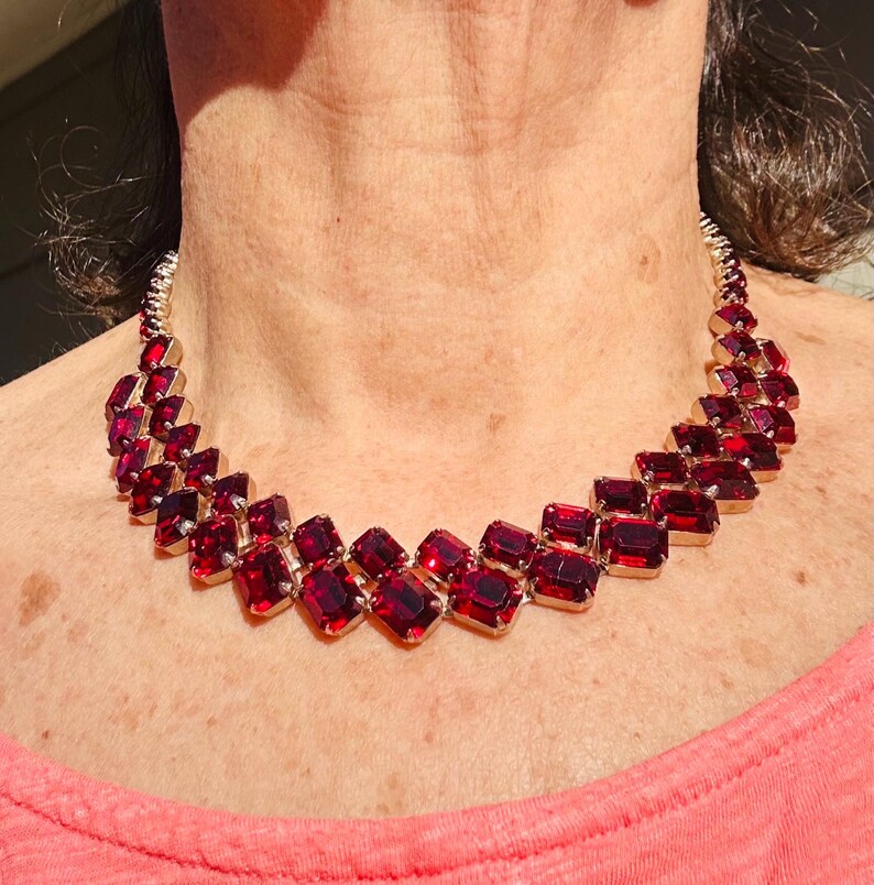 May include: A gold-coloured necklace with a series of rectangular, deep red gemstones. The stones are set closely together, creating a bold design. The necklace is a statement piece, ideal for adding a touch of glamour to an outfit.