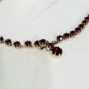 May include: A silver-toned necklace adorned with a sequence of round, deep red gemstones. The necklace showcases a classic design, with a single, larger gemstone pendant. The gemstones are evenly spaced along the chain.